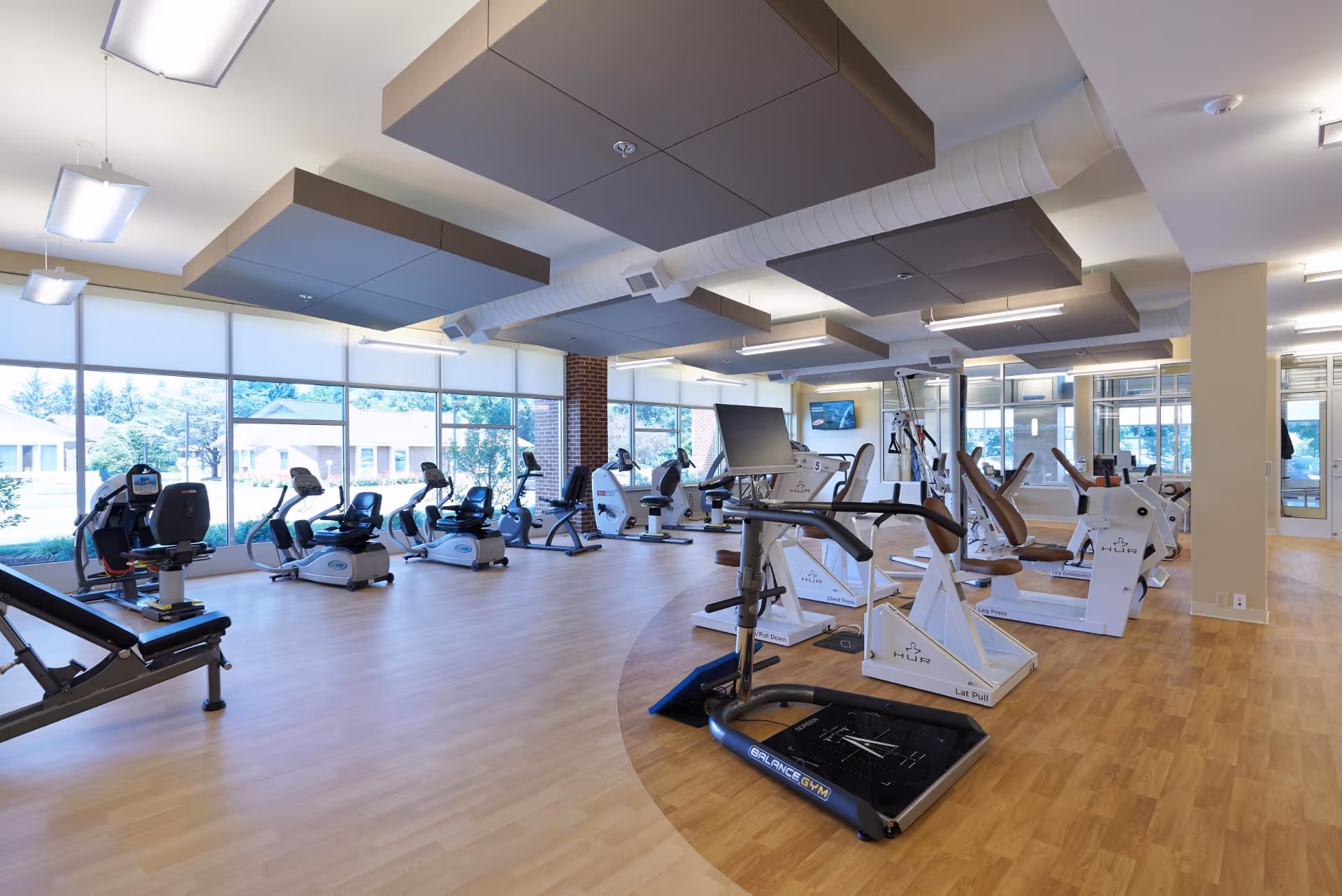 A spacious fitness room with large windows letting in natural light. The room contains various exercise equipment including stationary bikes, weight machines, and a balance gym platform. The floor is wood, and the ceiling has modern acoustic panels and lighting fixtures.
