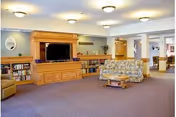 Spacious living room area in a nursing home with a large wooden entertainment center holding a flat-screen TV, built-in bookshelves filled with books, a floral patterned sofa, a wooden coffee table, and beige walls with ceiling lights.