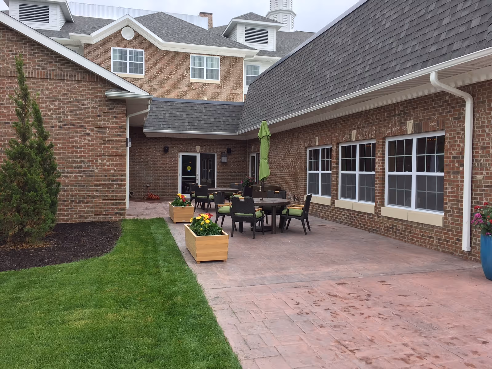 Outdoor patio area at Towne Center Retirement Community with brick walls, several windows, a green umbrella over a round table, and chairs with green cushions. There are wooden planters with flowers and a small grassy area adjacent to the patio.