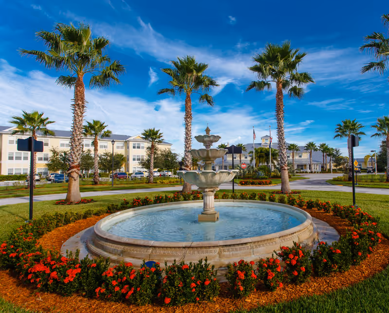 A multi-tiered decorative fountain surrounded by flowers and palm trees with a senior living building and entrance in the background under a blue sky.