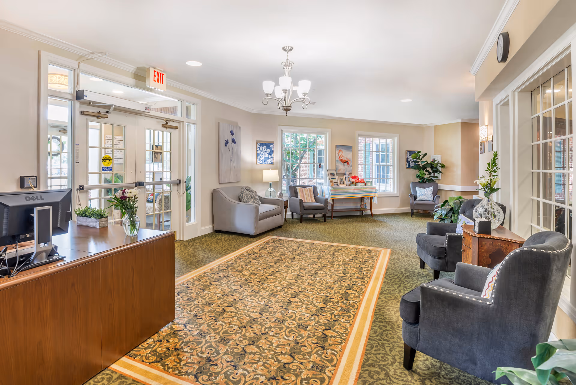 A bright and welcoming senior living facility lobby with a patterned carpet, comfortable armchairs, a small sofa, a wooden reception desk with a computer, and decorative plants and artwork on the walls. Large windows let in natural light, and there is an exit door with glass panels.