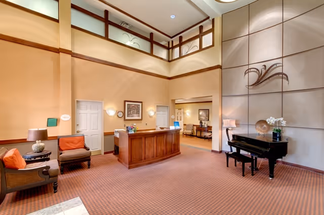 Bright, high-ceilinged reception lobby with a wooden front desk, seating area, decorative wall art, and a piano.
