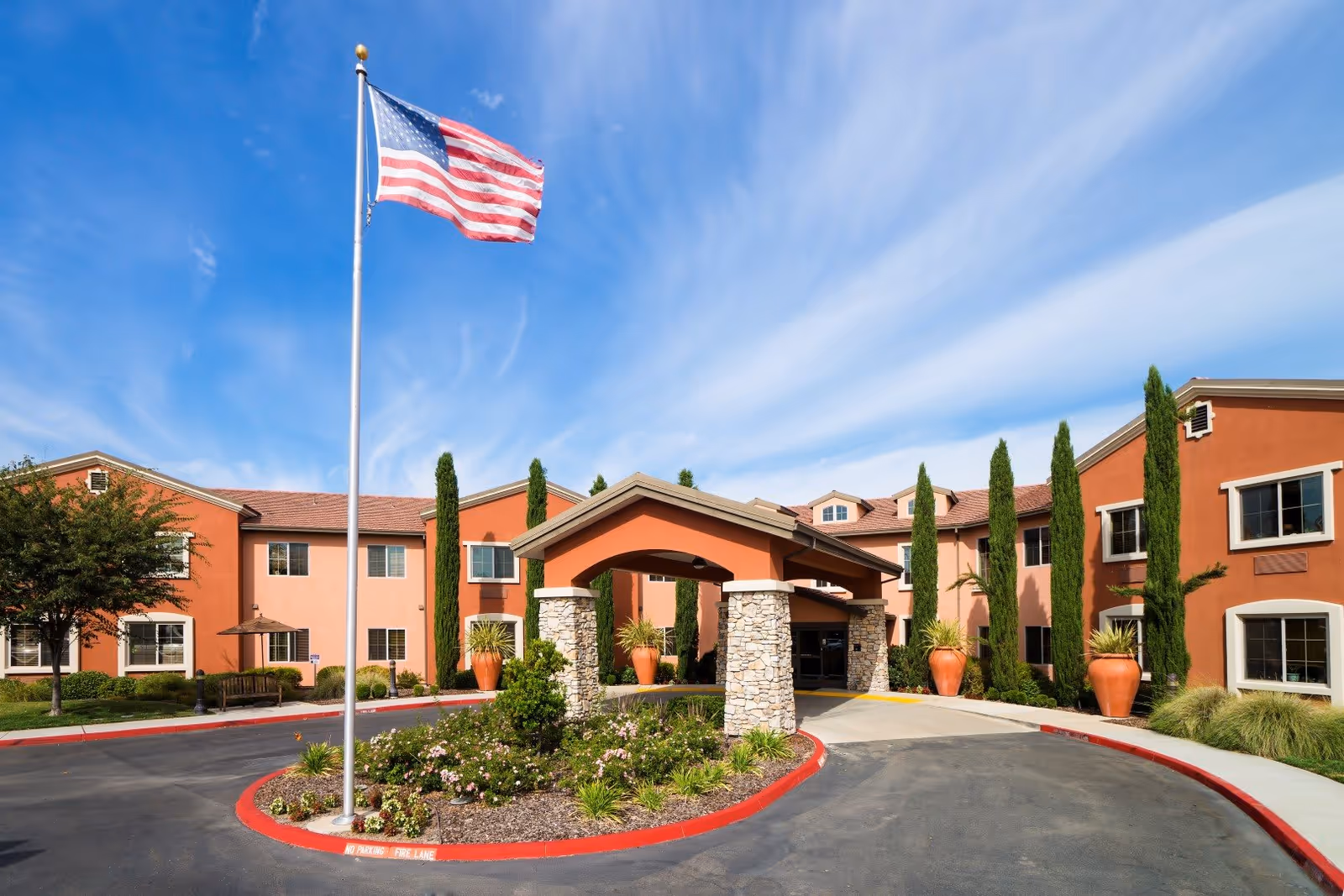 Exterior view of Eskaton Village Roseville building with a covered entrance supported by stone pillars, landscaped garden with flowers and shrubs, tall cypress trees, and an American flag flying on a flagpole against a blue sky with wispy clouds.
