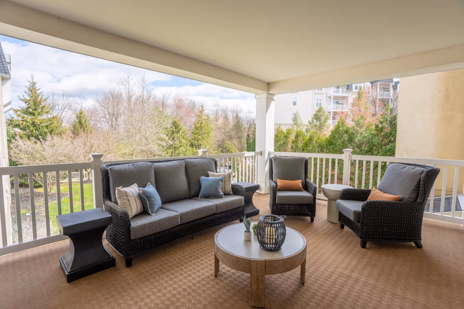 Covered outdoor patio area with wicker furniture including a sofa and two armchairs with cushions and pillows, a round wooden coffee table with decorative items, and side tables. The patio overlooks a landscaped garden with trees and shrubs.