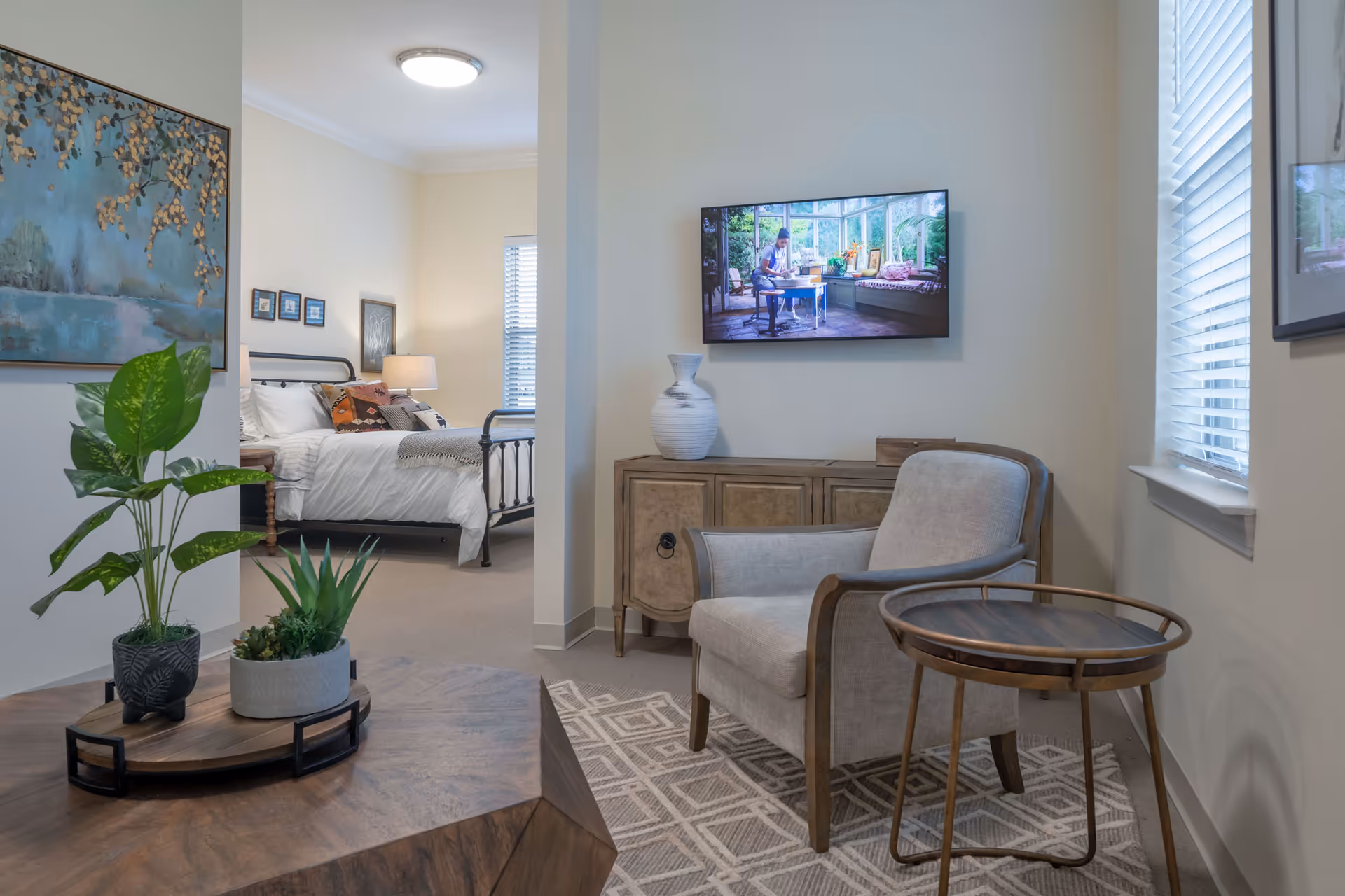 A cozy living area with a beige armchair and a round wooden side table on a patterned rug. A wooden cabinet with a decorative vase is against the wall, above which a flat-screen TV is mounted. The TV displays a person sitting at a table in a sunlit room. To the left, an open doorway reveals a bedroom with a neatly made bed, bedside lamp, and framed artwork on the walls. Two potted plants sit on a wooden coffee table in the foreground.