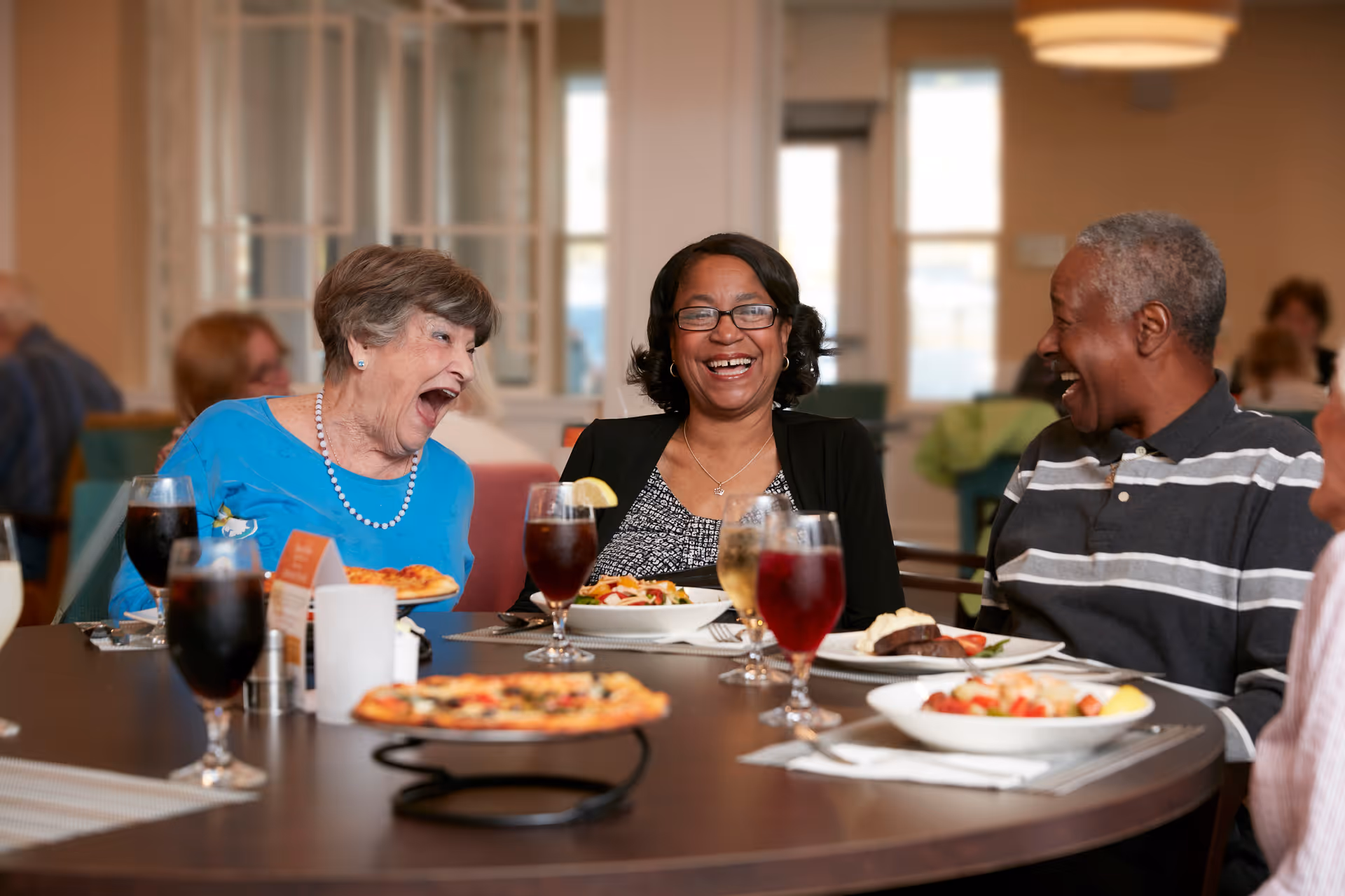 Three elderly people sitting around a dining table in a senior living facility, laughing and enjoying a meal together. The table has plates of food including pizza, salad, and drinks. The background shows a bright, warmly lit room with windows and other people dining.