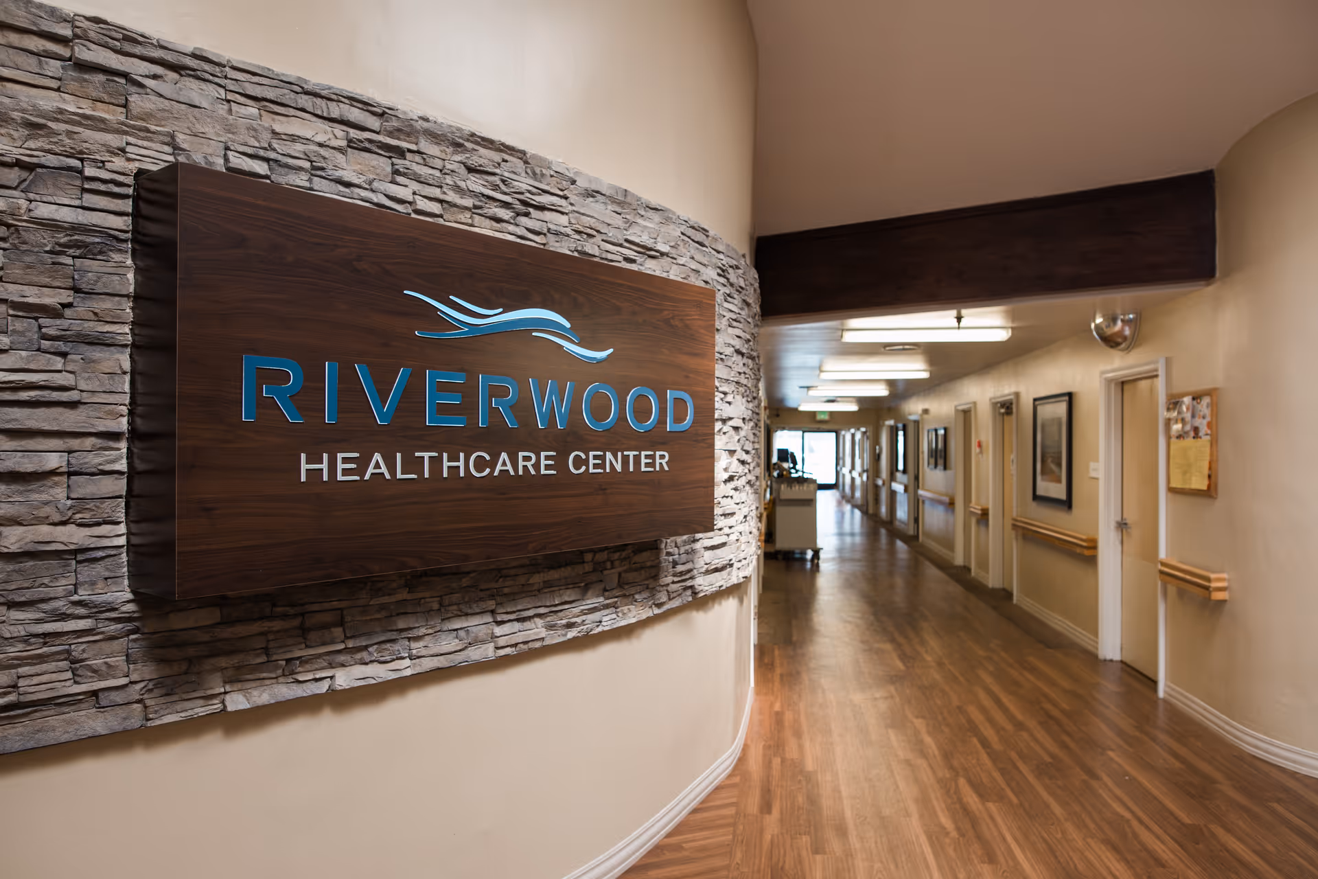 Interior hallway of Riverwood Healthcare Center featuring a large wooden sign on a stone wall and a long corridor with doors and wood flooring.