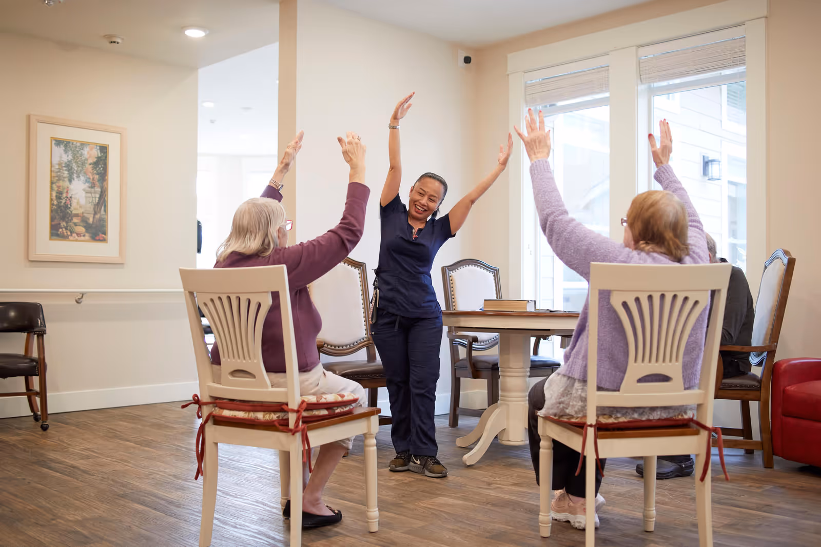 A group of elderly women seated on chairs in a bright room participate in a seated exercise session led by a smiling instructor who is standing with arms raised. The room has large windows, wooden flooring, and a table with chairs in the background.
