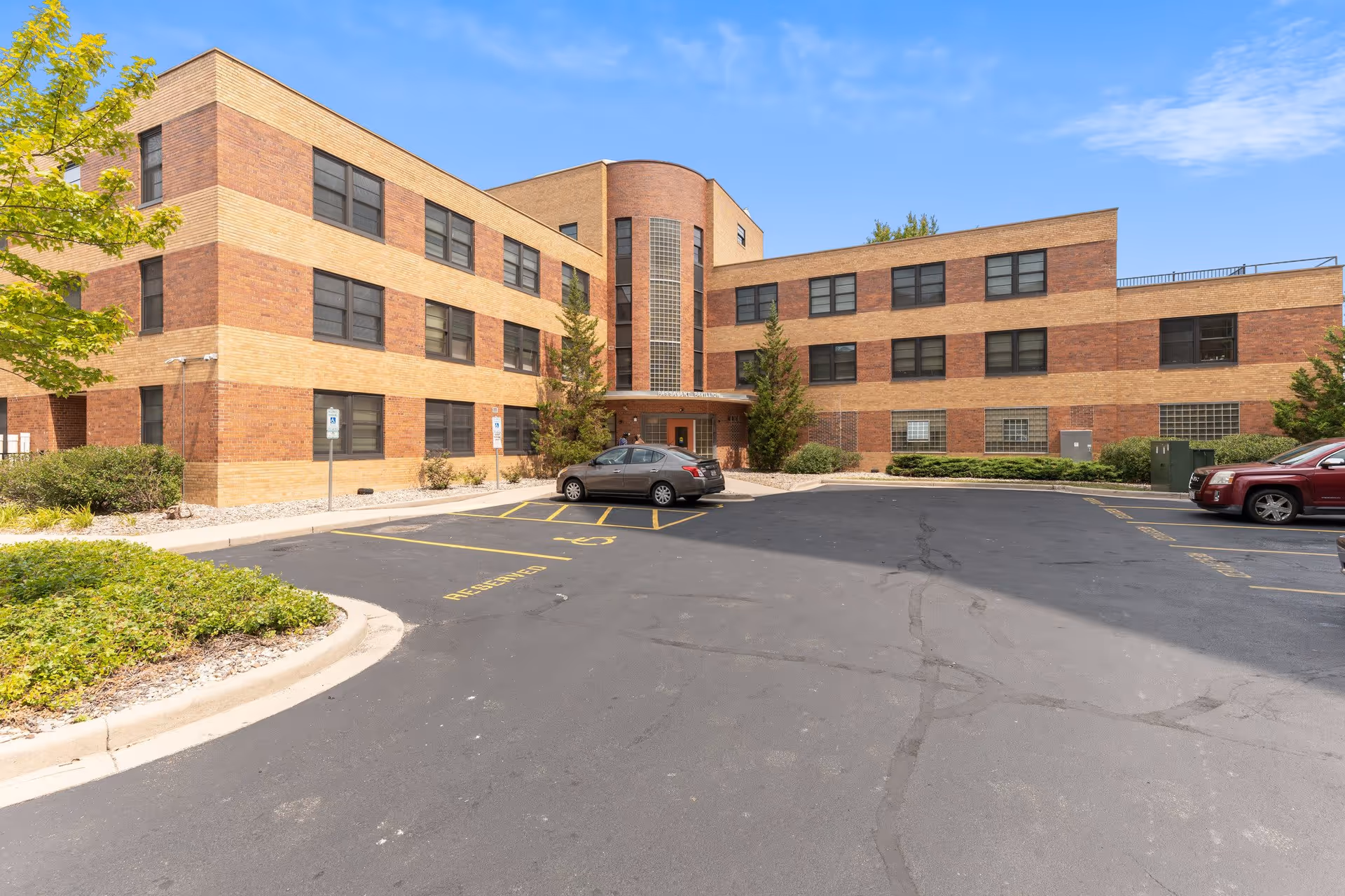 Exterior view of a three-story brick building with a parking lot in front. The building has multiple windows and a central entrance with a curved glass block feature above it. There are a few cars parked in the lot and some greenery around the building under a partly cloudy blue sky.