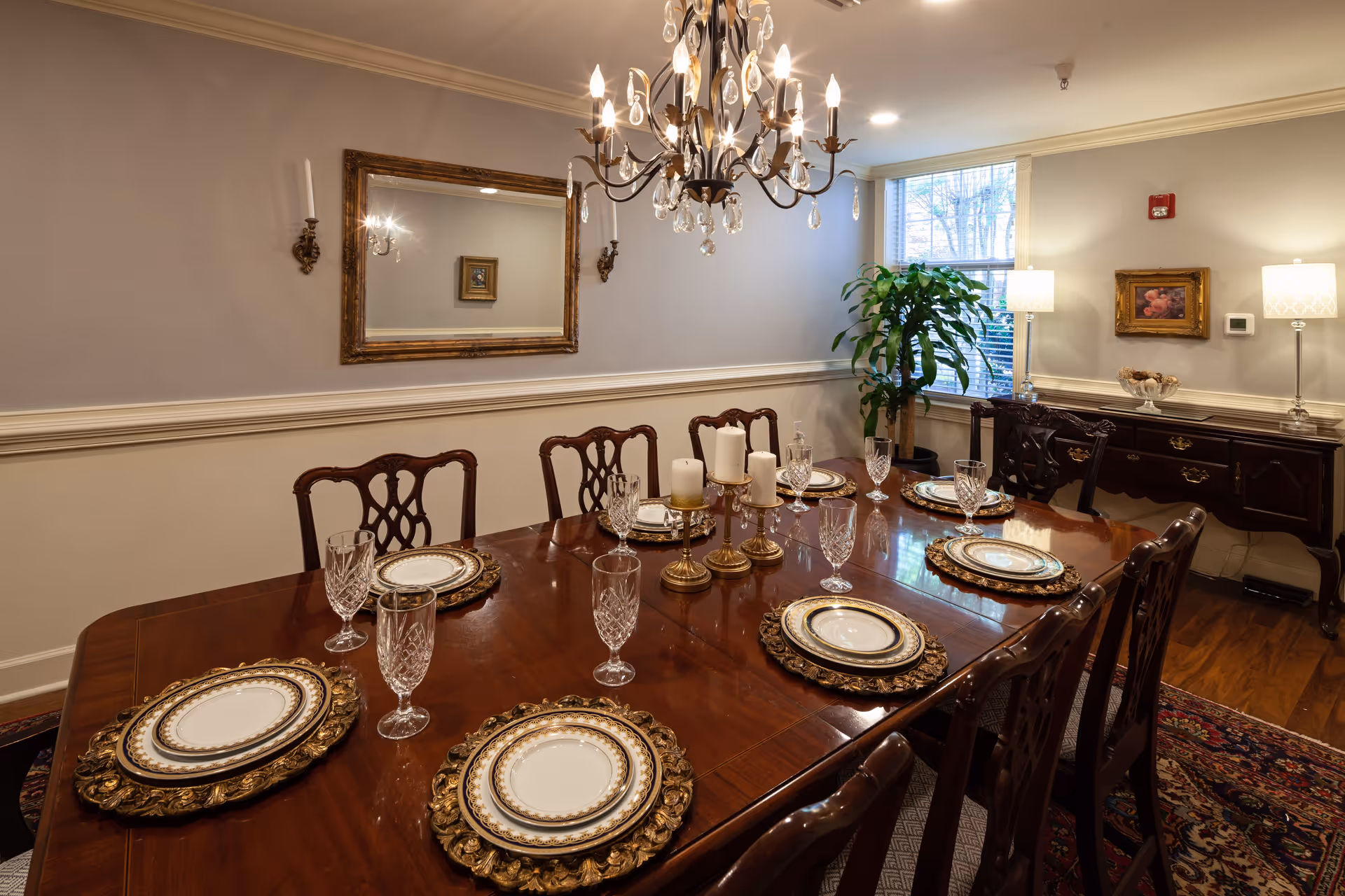 A formal dining room with a polished wooden table set for six people with ornate plates and crystal glasses. A chandelier with lit candles hangs above the table. The room has beige walls with white trim, a large mirror on one wall, two wall sconces, a window with natural light, a potted plant, and a dark wooden sideboard with two lamps and a framed picture above it.