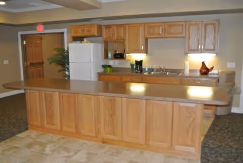 A kitchen area in a senior living facility featuring wooden cabinets, a white refrigerator, a coffee maker, a sink, and a long wooden island countertop. The room has beige walls and a mix of carpet and tile flooring.