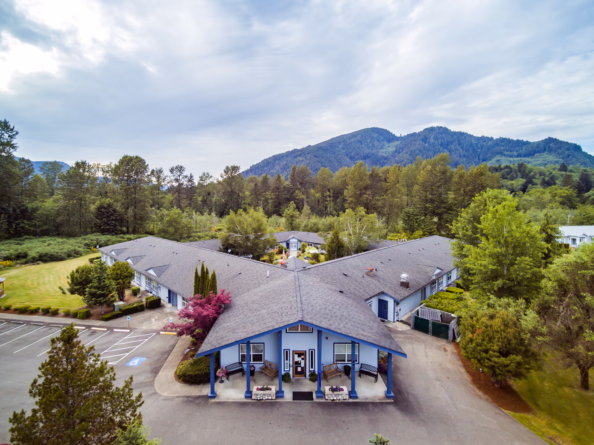 Aerial view of Heritage House Morton Assisted Living and Memory Care facility showing a single-story building with a Y-shaped layout surrounded by trees and greenery, with mountains in the background and a partly cloudy sky.