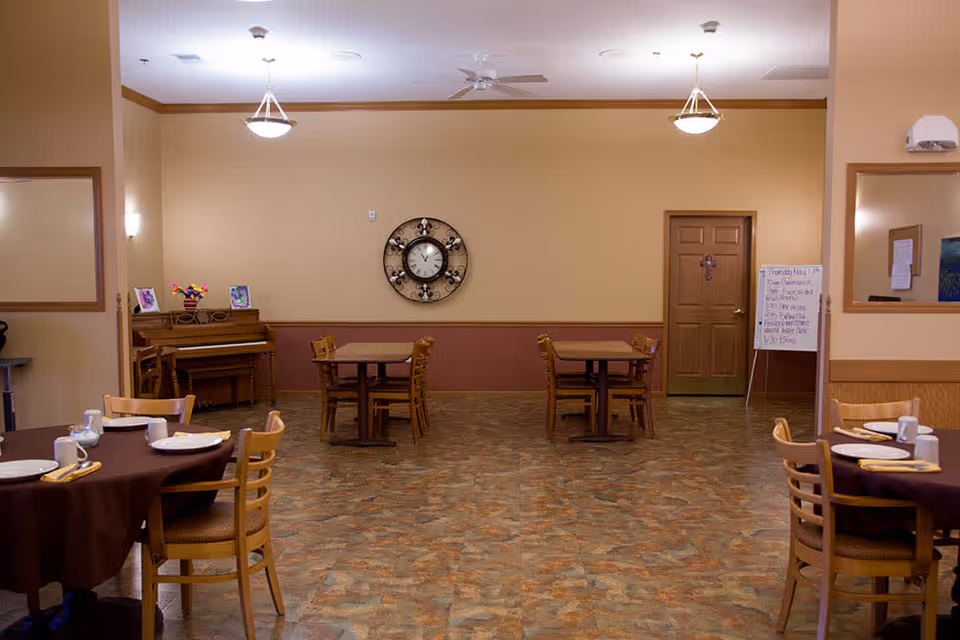 Interior view of a dining room in a senior living facility with several wooden tables and chairs arranged neatly. Two tables in the foreground are set with plates, cups, and utensils. A piano with framed pictures and a flower vase is against the left wall. A large decorative clock hangs on the back wall above two small tables with chairs. There is a door on the right side and a whiteboard with notes next to it. The room has warm beige walls, ceiling lights, and a patterned floor.