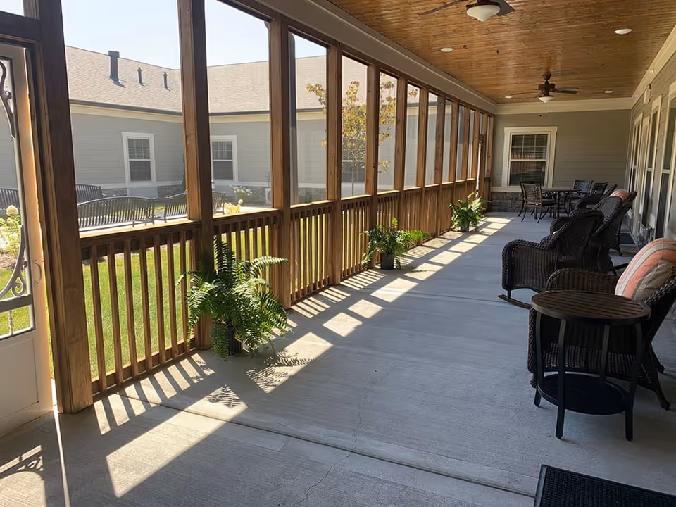 Shaded screened porch with wicker seating, small tables, potted ferns, and ceiling fans overlooking a grassy courtyard.