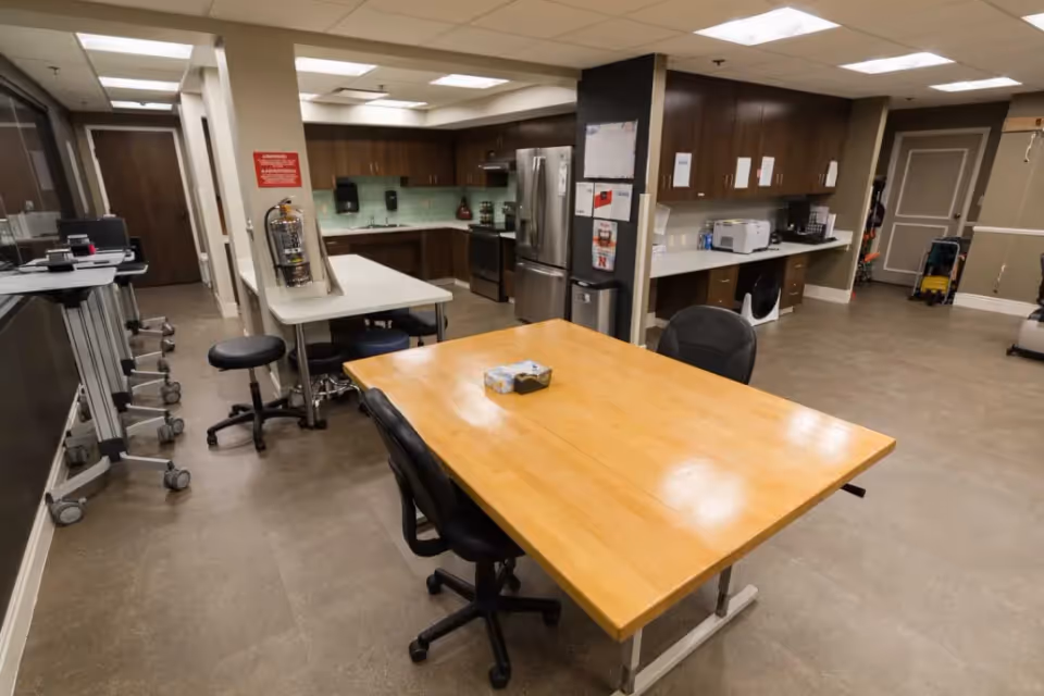 Interior view of a nursing facility kitchen and work area with a large wooden table surrounded by black office chairs in the foreground. The background shows a kitchen with stainless steel refrigerator, stove, microwave, and wooden cabinets. There are additional work tables, stools, and various office and kitchen equipment visible.