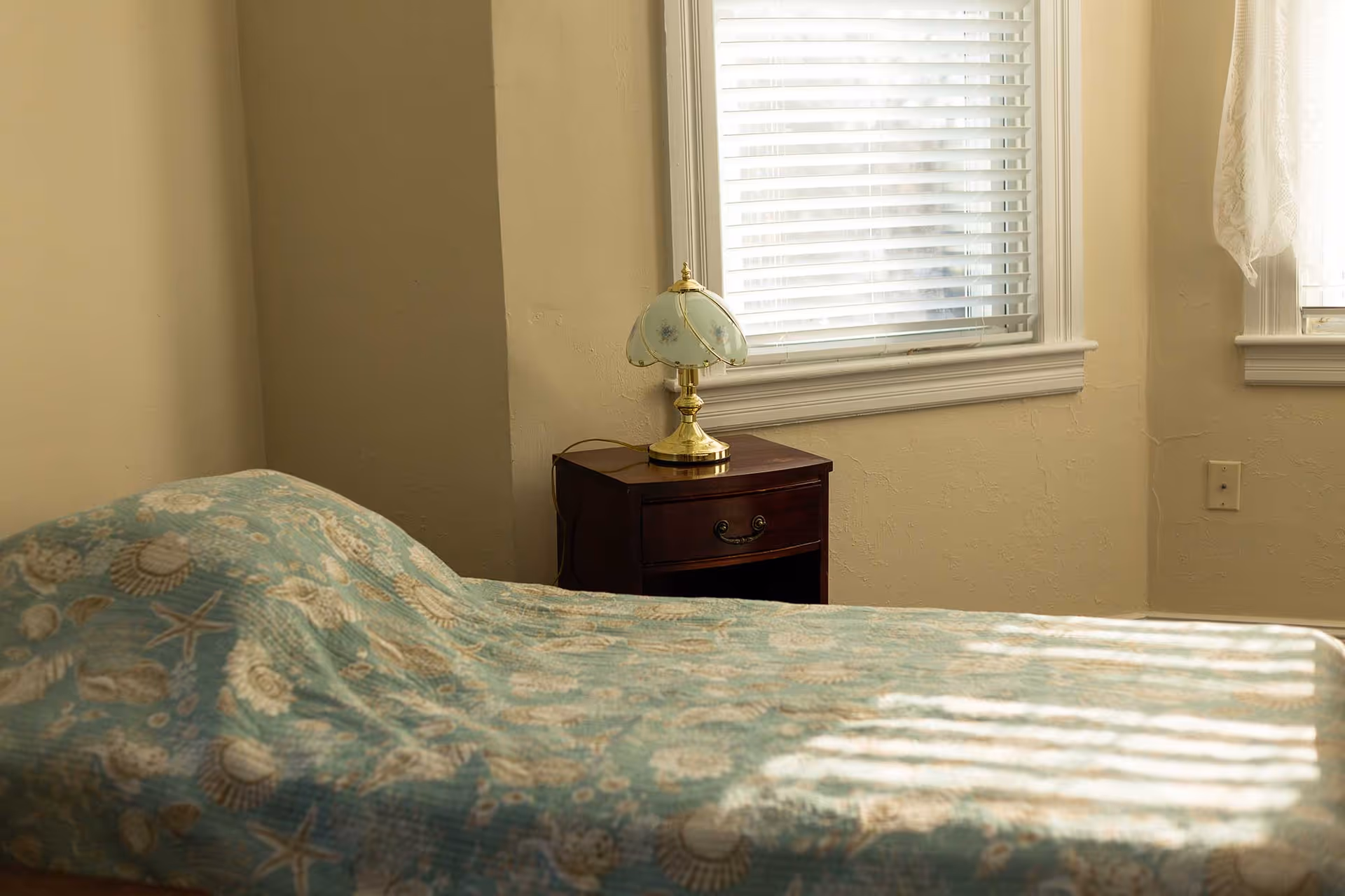 A small bedroom with a bed covered in a seashell-patterned quilt, a wooden nightstand and a decorative lamp beside a window with blinds.