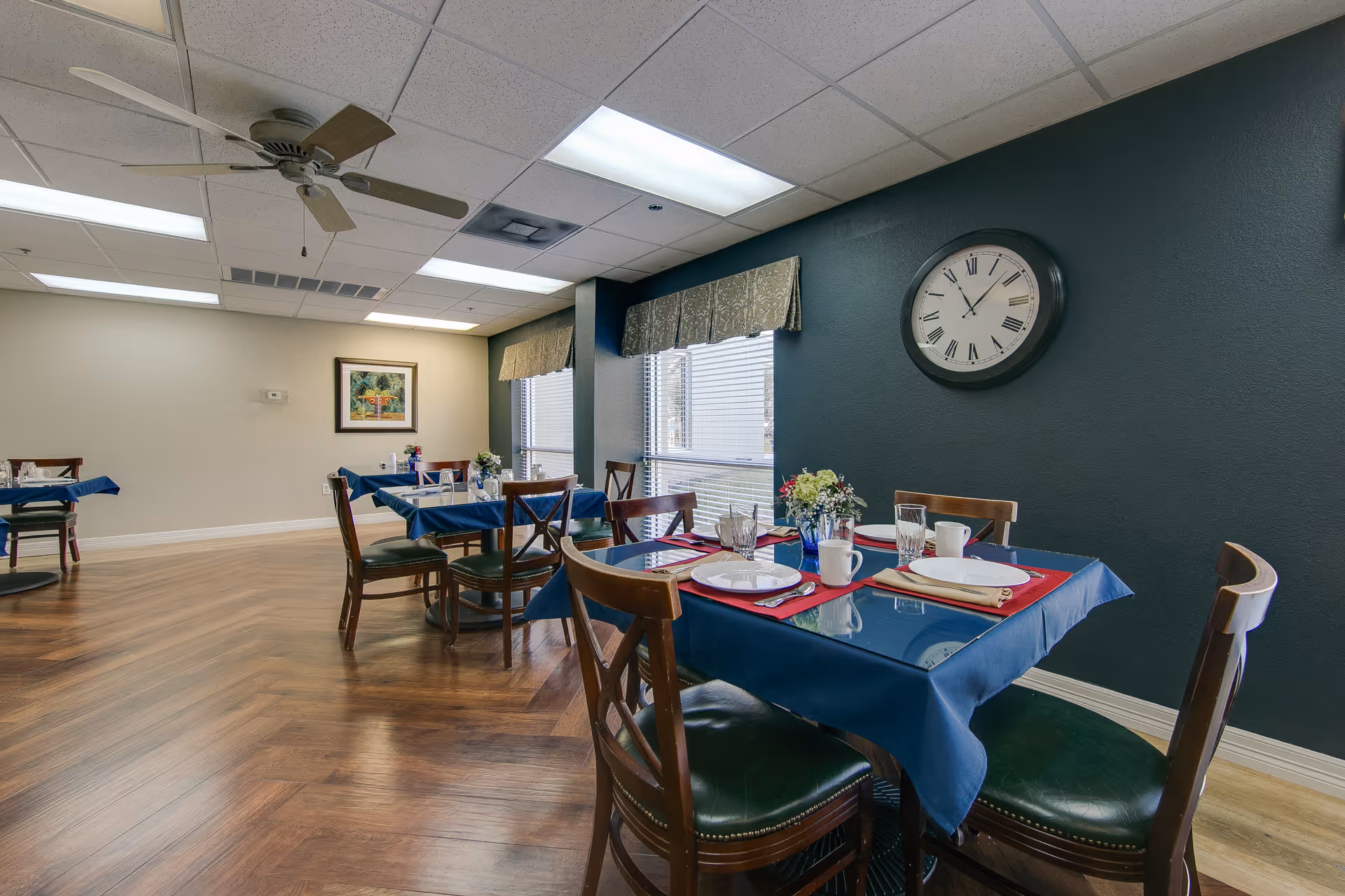 Dining room with wooden floors and several tables covered with blue tablecloths and set with plates, glasses, and napkins. There are wooden chairs with green cushions around the tables. A large clock with Roman numerals hangs on a dark green wall, and windows with valances let in natural light. A ceiling fan and fluorescent lights are visible on the ceiling.