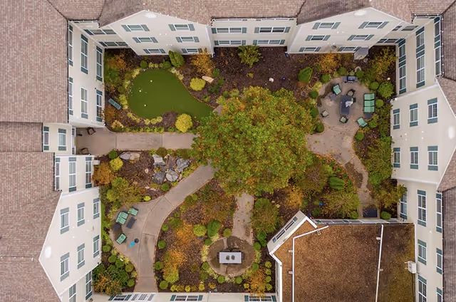 Aerial view of a courtyard garden surrounded by a multi-story residential building. The courtyard features a large tree in the center, walking paths, seating areas with chairs and tables, landscaped greenery, and a small putting green.