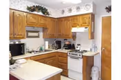 A compact kitchen with wooden cabinets, a small white stove, a microwave, a coffee maker, and various kitchen items on the countertops. The walls have a floral wallpaper border near the ceiling, and decorative plates are displayed above the cabinets.