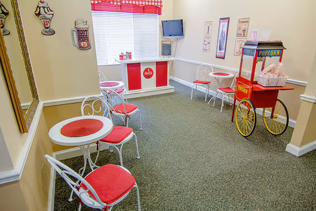 A small retro-style dining area with white round tables and white metal chairs with red cushions. There is a red and white popcorn cart on the right side and a small counter with a Coca-Cola logo under a window with red and white checkered curtains. The walls are decorated with vintage soda-themed signs.