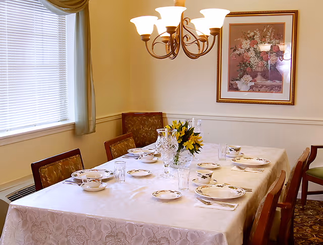 A formal dining room with a table set with china, crystal glassware and a yellow floral centerpiece under a chandelier.