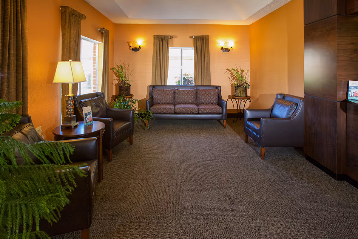 A cozy sitting room with leather chairs, a patterned sofa, side tables, lamps, and plants against warm orange walls.