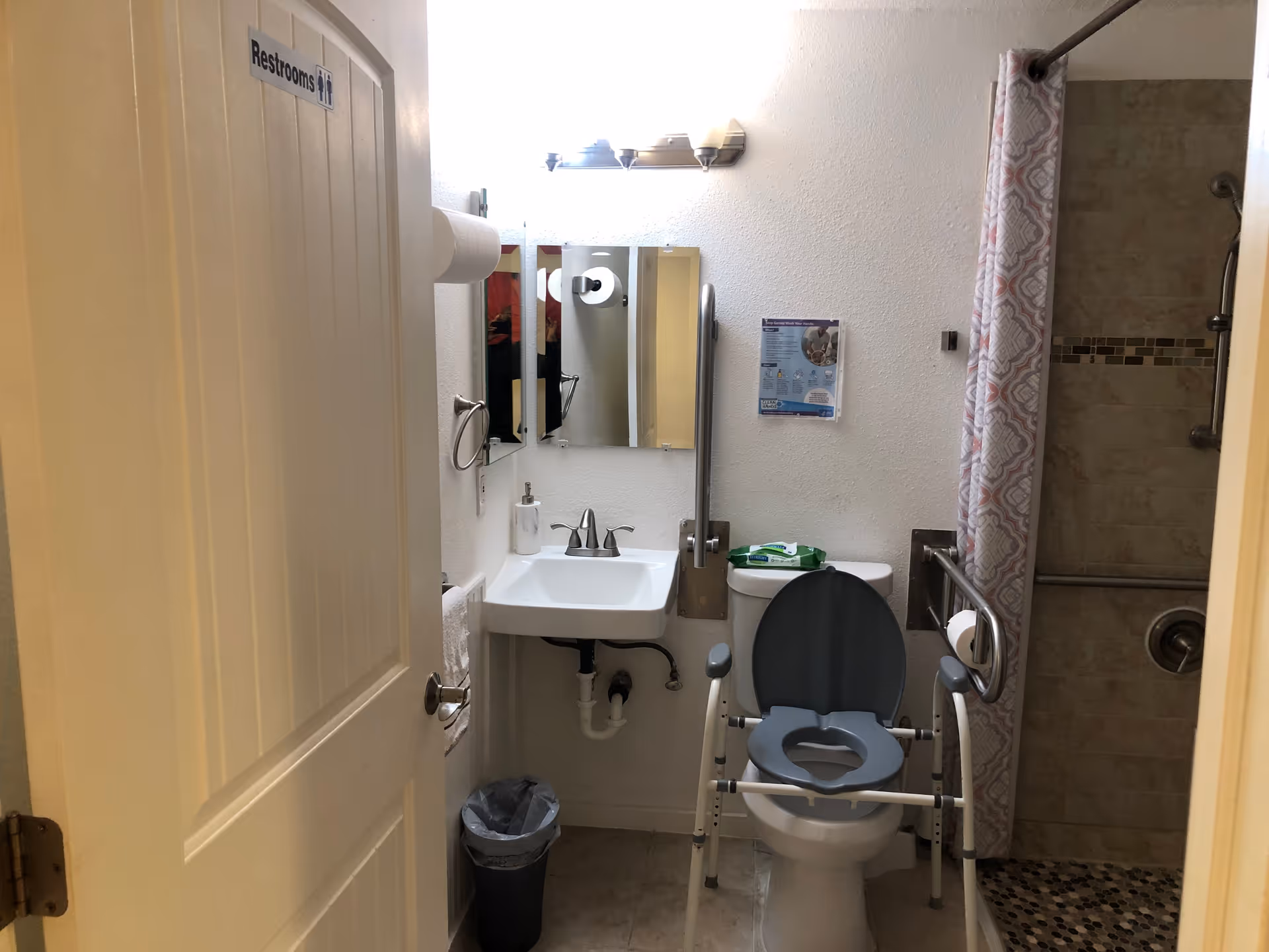 Small accessible bathroom with a toilet fitted with a safety frame, sink and mirror, and a walk-in shower with curtain, door labeled "Restrooms".