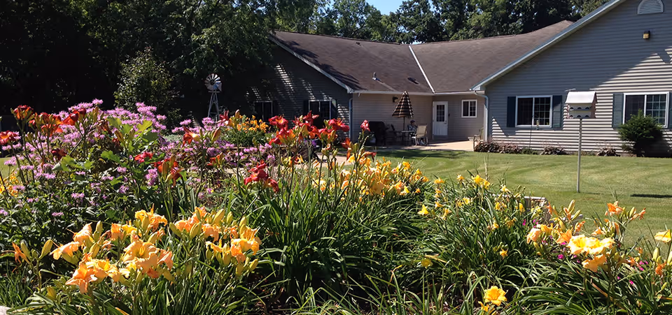 A garden with colorful flowers including yellow, orange, red, and purple blooms in front of a single-story building with beige siding and a brown roof. There is a small patio area with outdoor furniture and a birdhouse on a pole in the grassy yard.