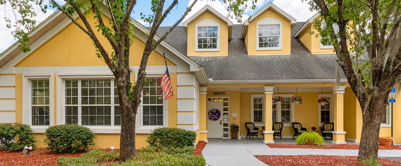 Front exterior view of a yellow building with white trim, featuring a covered porch with chairs and hanging flower baskets. Two trees and bushes are in the landscaped area with red mulch. An American flag is mounted near the window, and the building number 10875 is visible above the door.