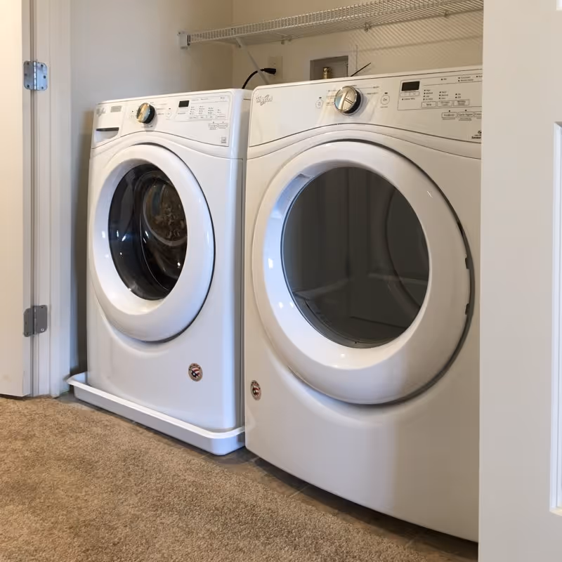 A laundry area with a white front-loading washing machine and a matching white front-loading dryer side by side on a carpeted floor. There is a wire shelf mounted on the wall above the machines.