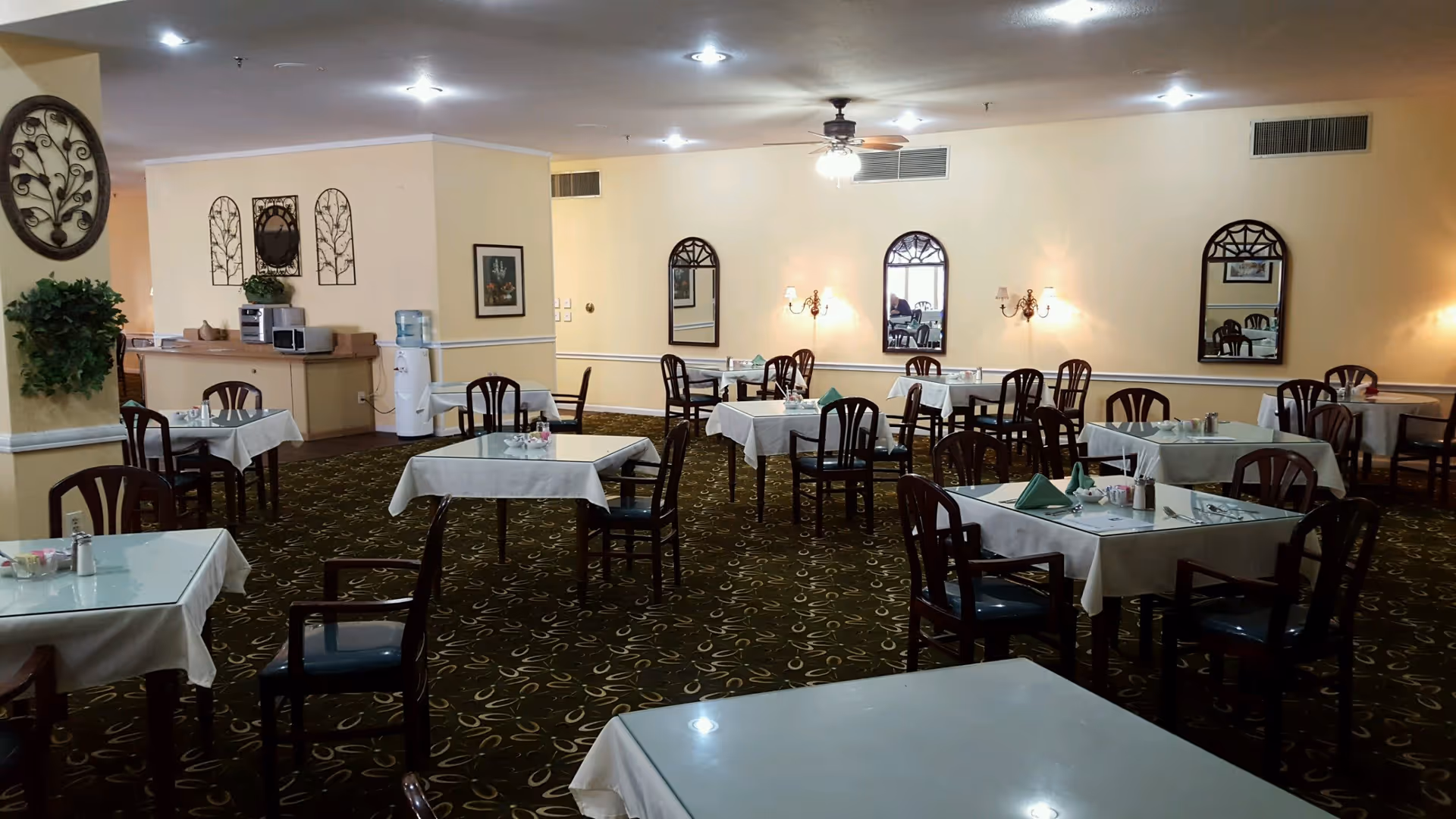 A dining room with multiple tables covered with white tablecloths and set with napkins, salt and pepper shakers, and small condiment containers. The room has patterned carpet, cream-colored walls with decorative mirrors and wall sconces, and a ceiling fan with lights. There is a water cooler and some kitchen appliances on a counter in the background.