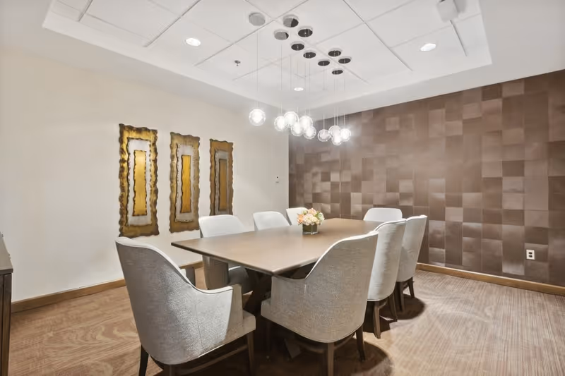 A modern dining room with a rectangular wooden table surrounded by eight upholstered chairs. A contemporary chandelier with multiple hanging glass globes is suspended above the table. The room features a textured brown accent wall and three decorative framed art pieces on a white wall. The floor is carpeted with a subtle pattern.