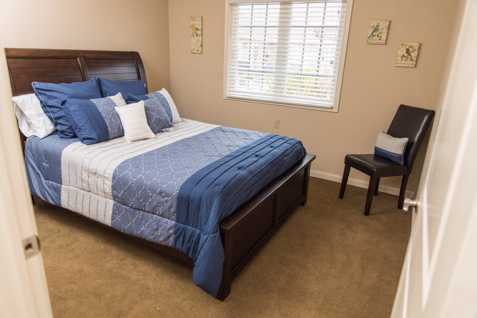 A bedroom with a wooden bed frame and a neatly made bed featuring blue and white bedding and multiple pillows. There is a window with white blinds on the wall behind the bed, two small bird-themed wall decorations, and a dark brown chair with a cushion in the corner of the room. The carpet is beige and the walls are painted a light beige color.