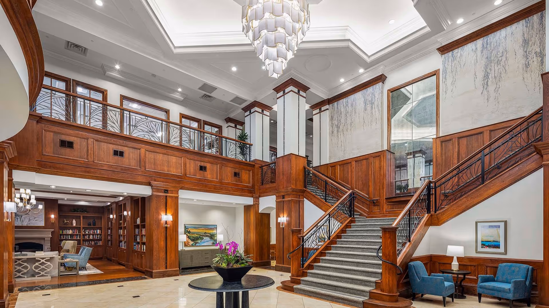 Spacious and elegant interior of a senior living facility featuring a grand staircase with wooden railings and paneling, a large chandelier hanging from the ceiling, comfortable blue armchairs with a small table and lamp, and a cozy library area with bookshelves and seating in the background.