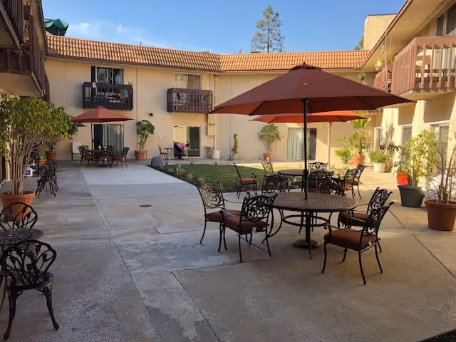 Outdoor courtyard area of a retirement facility with several round metal tables and chairs, each table shaded by a large red umbrella. The courtyard is surrounded by a two-story building with balconies and potted plants placed around the seating area. The sky is clear and blue.