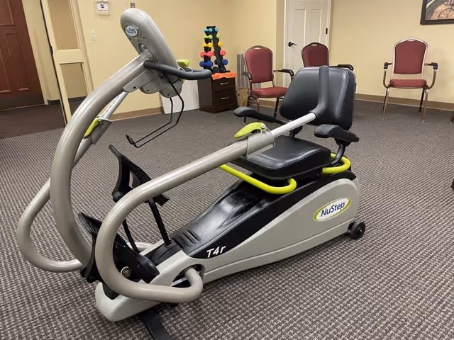 A NuStep T4r recumbent cross trainer exercise machine placed on a carpeted floor in a room with beige walls. In the background, there are three red cushioned chairs, a small set of colorful dumbbells on a stand, and a closed white door.