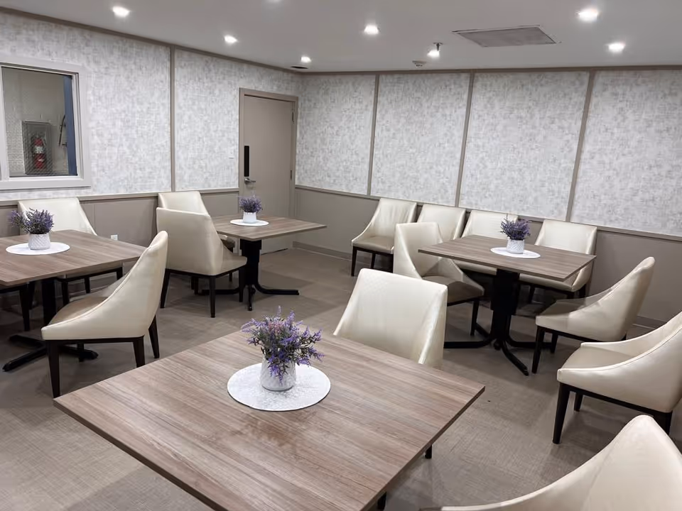 A small dining area with several square wooden tables, each adorned with a small white vase containing purple flowers. The room has beige cushioned chairs around the tables, light-colored walls with a textured pattern, and a door in the back. The lighting is bright with recessed ceiling lights.