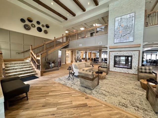 Spacious senior living facility common area with wooden flooring, a large rug, comfortable armchairs, a couch, and a modern fireplace with a map artwork above it. There is a staircase with wooden railings leading to an upper level, and decorative wall art on the left wall. The area is well-lit with natural light coming from windows in the background.
