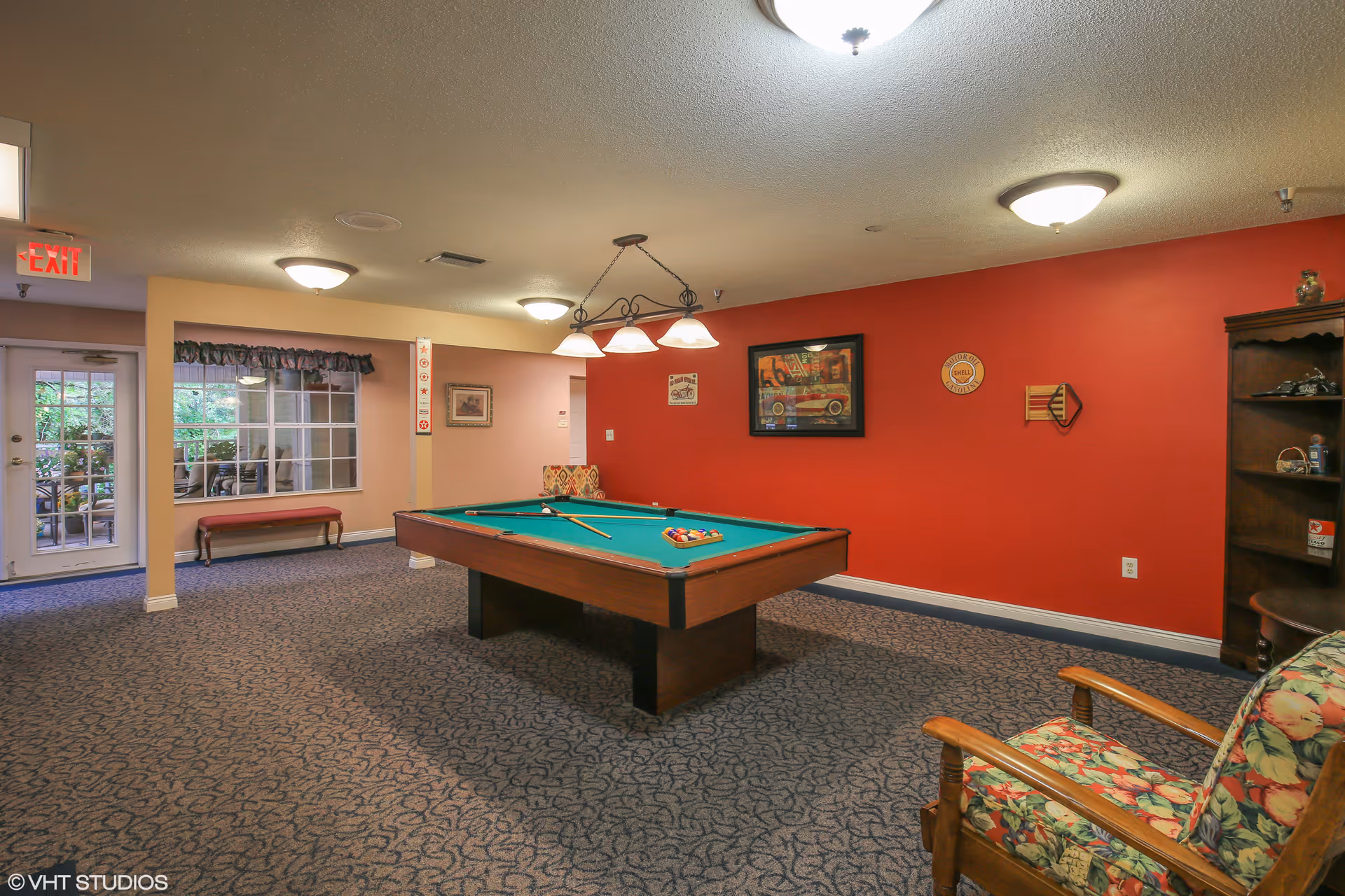 A recreation room with a pool table centered under pendant lights, a red accent wall, seating and shelving.