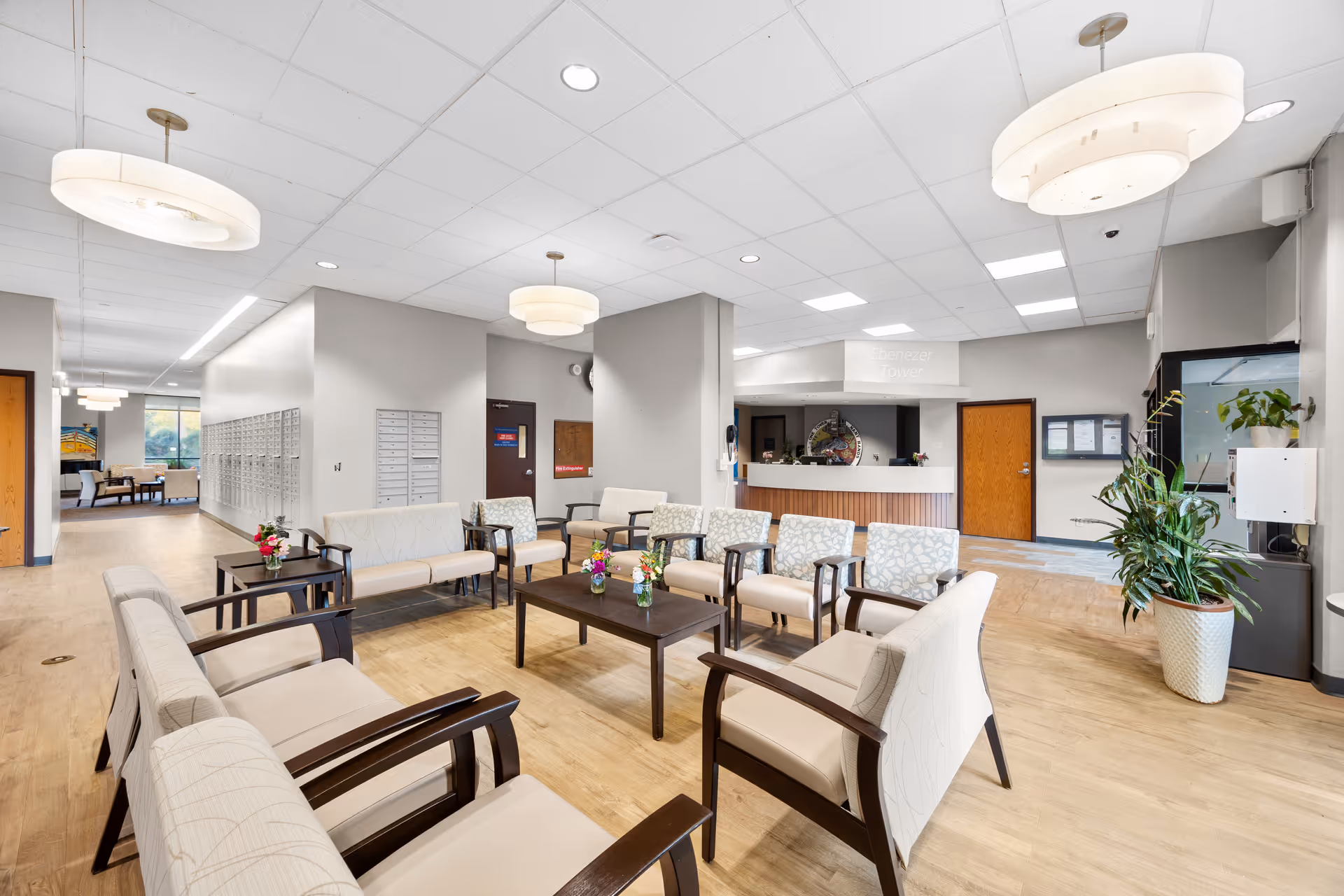 A bright and spacious senior living facility lobby with multiple beige cushioned chairs and sofas arranged around dark wooden coffee tables with small flower vases. The room has light wood flooring, white ceiling tiles with recessed lighting, and modern circular pendant lights. In the background, there is a reception desk with a sign above it that reads 'Ebenezer Tower'. Mailboxes line one wall, and there are large windows letting in natural light. A large potted plant is placed near the entrance area.