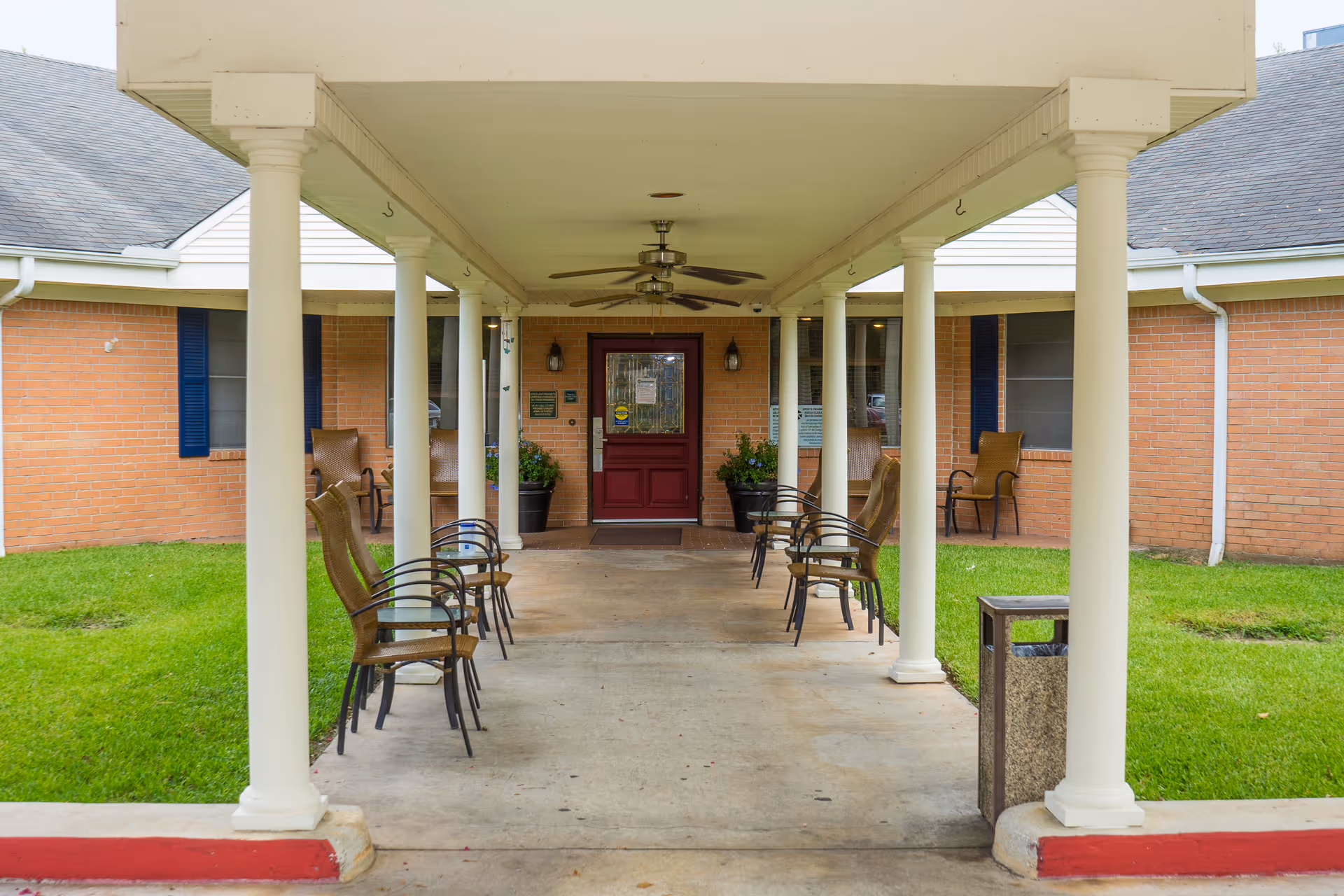 Covered walkway with white columns and ceiling fans leading to a red door entrance of a brick building. Several chairs are arranged along the walkway on both sides, with green grass on either side of the path.