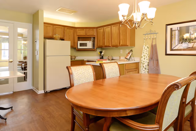 A kitchen and dining area with wooden cabinets, a white refrigerator, a microwave, and a wooden dining table with six upholstered chairs. The walls are painted light green, and there is a chandelier above the table. A framed picture and hanging aprons are visible on the wall.