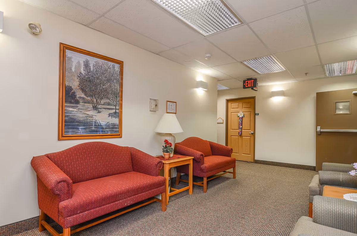 A waiting area with two red upholstered sofas and a wooden side table with a lamp and a small potted plant. There is a framed picture of a tree on the wall above one sofa. The area has beige walls, carpeted floor, and ceiling lights. A wooden door with a decorative hanging and an exit sign above it is visible in the background.