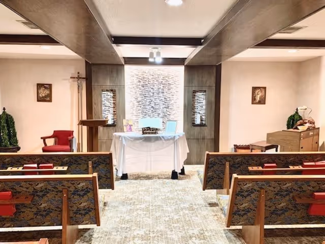 Interior view of a small chapel or prayer room with wooden pews facing a white altar covered with a cloth. The altar is set against a textured wall with soft lighting. There is a wooden lectern to the left, a cross on the wall, framed religious pictures, and some greenery decorations on either side.