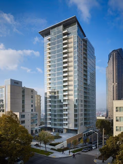 Tall modern residential building with glass balconies and large windows surrounded by other buildings and trees under a partly cloudy blue sky.