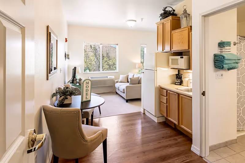 Interior view of a senior living facility model apartment at The Chesterley, showing a small kitchen area with wooden cabinets, a refrigerator, microwave, and coffee maker on the right. To the left, there is a round table with a chair and a 'MODEL' sign on the table. In the background, a living room area with a beige sofa, a window with a view of trees, and a TV on a stand is visible. A bathroom entrance with teal towels hanging on a rack is seen on the right side.