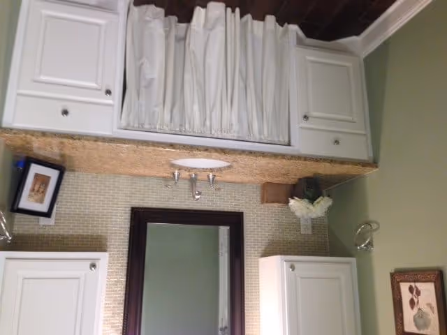 Bathroom with a white sink and countertop, white cabinets above and below the sink, a rectangular mirror with a dark brown frame, a small framed picture leaning against the wall, a vase with white flowers on the countertop, and a towel ring mounted on the wall next to the mirror.