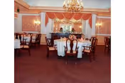 Elegant dining room with round tables covered in white tablecloths, wooden chairs, chandeliers, and decorative wall panels with warm lighting.