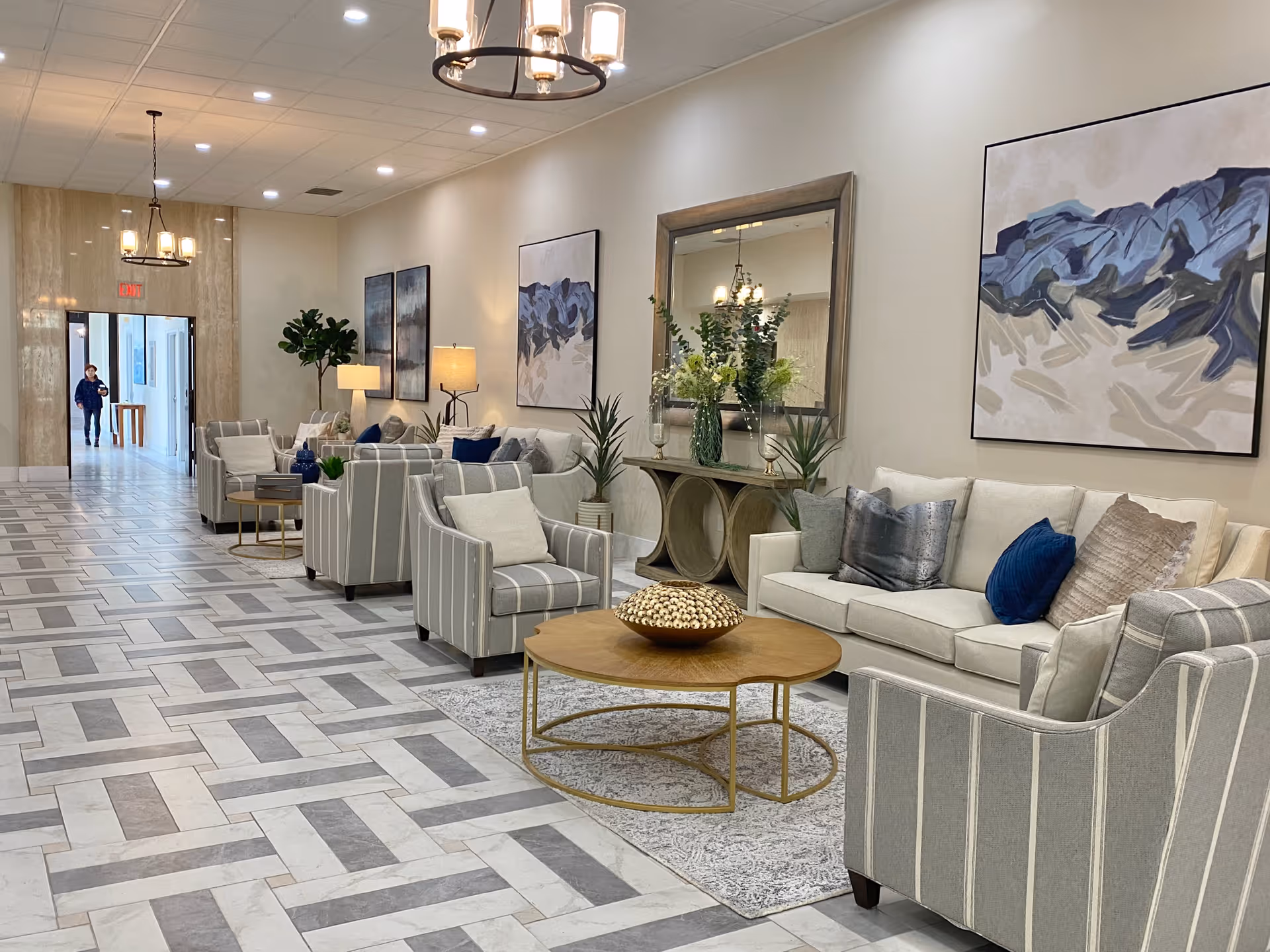 A spacious and well-lit senior living facility lounge area with a patterned tile floor, several striped armchairs, a white sofa with decorative pillows, a round wooden coffee table, and a console table with a large mirror and floral arrangements. Modern paintings hang on the walls, and a person is seen walking in the hallway in the background.