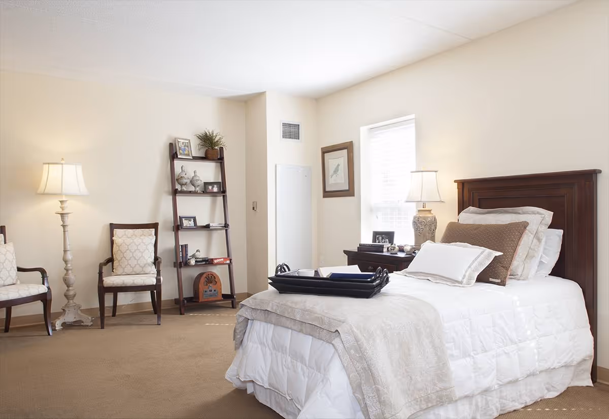 A bright and neatly arranged bedroom with a single bed featuring white and beige bedding and multiple pillows. There is a dark wooden headboard and a nightstand with a decorative lamp and picture frames next to the bed. Two upholstered chairs and a tall floor lamp are positioned against the wall. A leaning ladder-style shelf holds decorative items and books. The room has beige walls and carpet, with a window allowing natural light to enter.